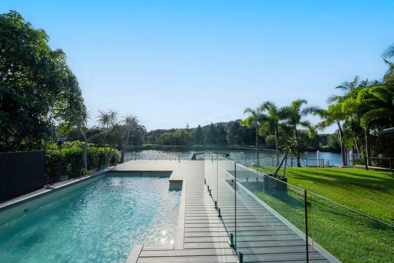 A pale composite pool deck with glass fencing, palms and a waterway beyond.